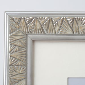 May include: A close-up of a silver picture frame with a detailed, textured design. The frame has a light grey colour with a decorative pattern in a lighter, beige tone. The frame surrounds a white mount, suggesting it is for displaying artwork or photographs.