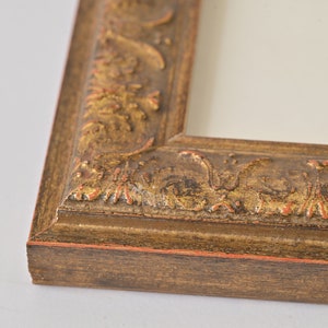 May include: A close-up of a gold picture frame with ornate detailing. The frame has a dark brown wood base and a gold-coloured, raised, floral design.