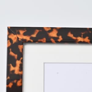 May include: A rectangular picture frame featuring a glossy tortoiseshell pattern with mottled brown and amber tones. The frame includes a clean white mat board border surrounding the central opening, showcasing a mitered corner joint construction.