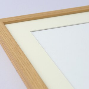 May include: A close-up of a rectangular wooden picture frame with a light beige mat and a white interior. The frame is made of light-colored wood and has a simple, clean design. The mat is a soft, off-white color.
