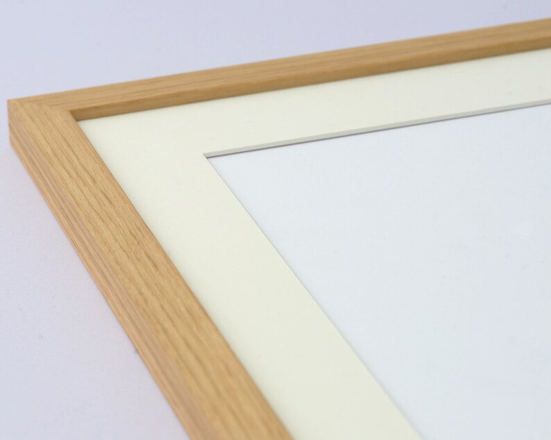 May include: A close-up of a rectangular wooden picture frame with a light beige mat and a white interior. The frame is made of light-colored wood and has a simple, clean design. The mat is a soft, off-white color.