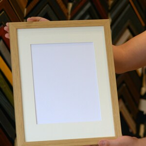 May include: A rectangular, light-colored wooden picture frame with a white mat and a blank white space for a photo or artwork. The frame is held by a person, showcasing the natural wood grain and the clean lines of the design. The background features a variety of other frames.