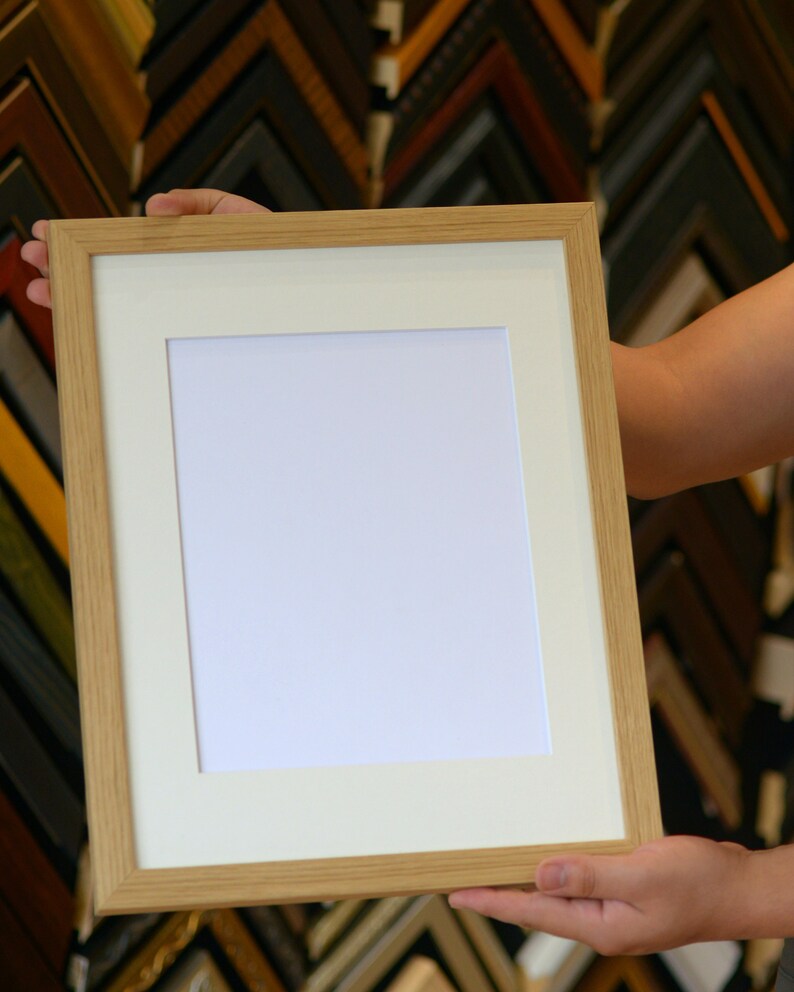 May include: A rectangular, light-colored wooden picture frame with a white mat and a blank white space for a photo or artwork. The frame is held by a person, showcasing the natural wood grain and the clean lines of the design. The background features a variety of other frames.