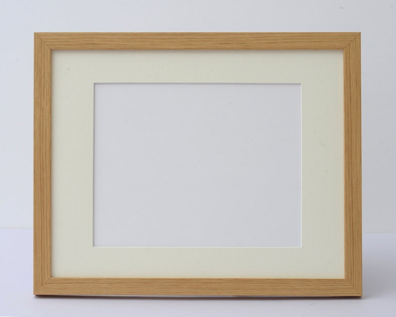 May include: A rectangular wooden picture frame with a light brown finish. The frame has a cream-colored mat and a white interior space for a photograph or artwork. The frame is designed for displaying images.