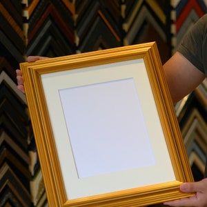May include: A gold picture frame with a white mat and a blank white space inside. The frame is being held by a person's hand.