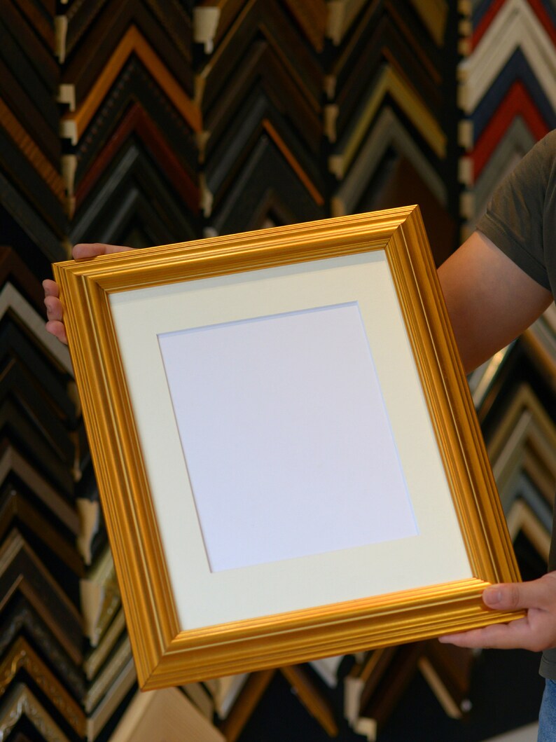 May include: A gold picture frame with a white mat and a blank white space inside. The frame is being held by a person's hand.