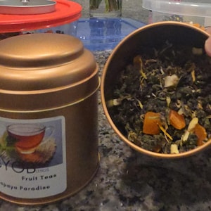 Puede incluir: Una lata de metal marrón con tapa dorada que contiene té de hojas sueltas. La lata tiene una etiqueta que dice "BYOB Loose Leaf Teas Fruit Teas Papaya Paradise". El té es una mezcla de té verde, papaya y otras frutas.