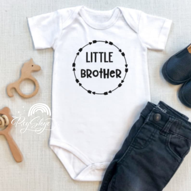 Puede incluir: Un body blanco para beb&eacute; con texto negro que dice "LITTLE BROTHER" dentro de un c&iacute;rculo de flechas negras.