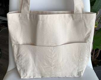 May include: A plain white canvas tote bag with a large front pocket. The bag has two long straps.