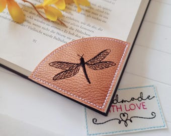 May include: A copper-colored leather bookmark with a black embroidered dragonfly design. The bookmark is placed on the corner of a book. A small tag with the words "handmade with LOVE" is also visible.