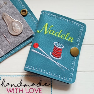 May include: A teal blue leather needle case with white stitching. The case has a gold button and is embroidered with the word "Nadeln" in green thread, a spool of red thread, and two white needles. The case is handmade and has a label that says "handmade with love".