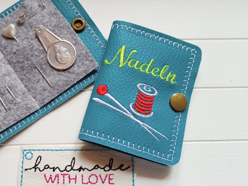 May include: A teal blue leather needle case with white stitching. The case has a gold button and is embroidered with the word "Nadeln" in green thread, a spool of red thread, and two white needles. The case is handmade and has a label that says "handmade with love".