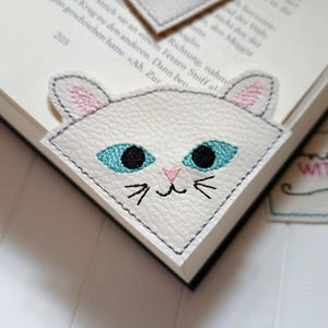 Könnte beinhalten: Weißer Leder-Lesezeichen mit einem gestickten Katzenkopf-Design. Die Katze hat blaue Augen, eine rosa Nase und weiße Schnurrhaare. Das Lesezeichen ist mit grauem Garn gestickt.
