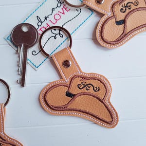 Könnte beinhalten: Zwei braune Leder-Schlüsselanhänger mit einem braunen gestickten Cowboyhut-Design. Die Schlüsselanhänger haben silberne Schlüsselringe und sind an einem silbernen Schlüssel befestigt.