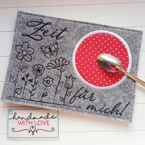 Könnte beinhalten: Grauer Filz-Platzdeckchen mit gestickten Blumen, Schmetterling und deutschem Text. Ein roter Kreis mit weißen Punkten und weißem Rand hält einen Löffel. Ein kleines Etikett lautet "handmade with love".