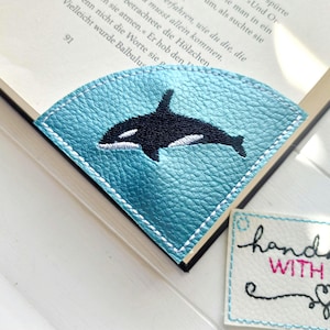 Könnte beinhalten: Ein türkisfarbener Leder-Lesezeichen mit einem schwarz-weißen Orca-Wal-Design. Das Lesezeichen ist mit weißem Faden genäht und befindet sich in einem Buch. Ein kleines Etikett mit dem Text "handmade WITH LOVE" ist ebenfalls sichtbar.