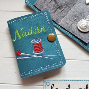 May include: A teal blue needle case with a white stitched border. The case has a brown button and embroidered text that says "Nadeln" in green. The case also has an embroidered image of a spool of red thread and two needles.
