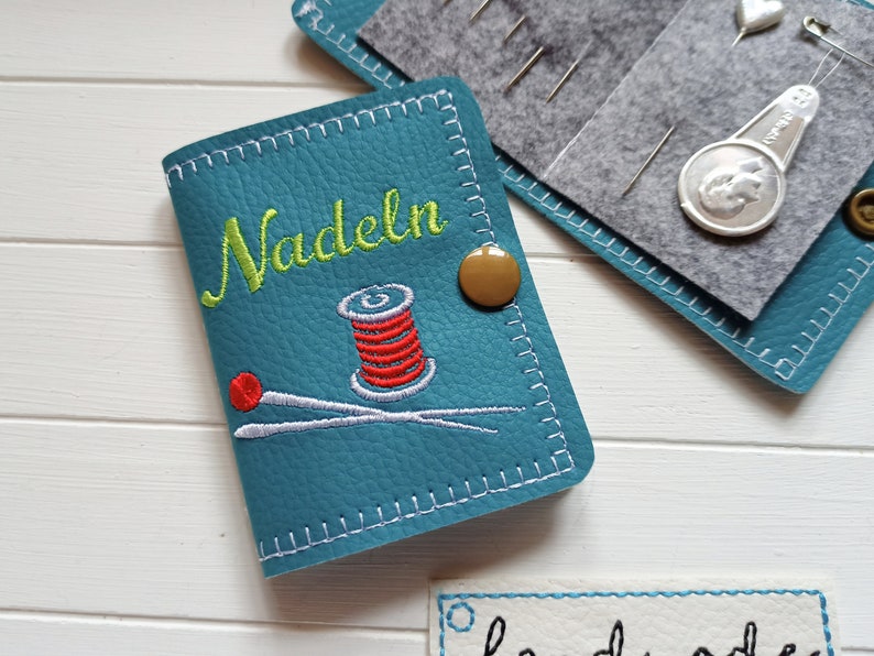 May include: A teal blue needle case with a white stitched border. The case has a brown button and embroidered text that says "Nadeln" in green. The case also has an embroidered image of a spool of red thread and two needles.