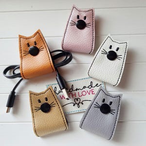 Könnte beinhalten: Eine Auswahl an kabelorganisierenden Katzen in verschiedenen Farben, darunter Beige, Rosa, Weiß und Grau. Jede Katze hat eine schwarze Knopf-Nase und gestickte Schnurrhaare. Ein Kabel ist an einem der Organizer befestigt. Der Schriftzug "handmade with LOVE" befindet sich auf einem Etikett.