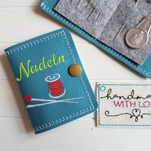 May include: A teal blue needle case with the word "Nadeln" embroidered on the front. The case features a small spool of thread and two needles embroidered on the front. The case has a gold button closure. A white tag with the text "handmade with love" is in the background.