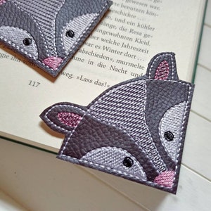 Könnte beinhalten: Ein graues und pinkfarbenes Waschbär-Lesezeichen mit gestickten Details. Das Lesezeichen ist in Form einer Buchseite geformt.