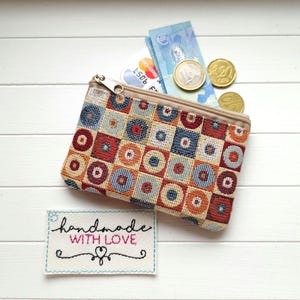 Könnte beinhalten: Eine kleine, rechteckige Geldbörse mit einem farbenfrohen geometrischen Muster in Rot, Blau, Orange und Beige. Die Geldbörse ist geöffnet und zeigt Bargeld, Münzen und Kreditkarten. Ein kleines Etikett lautet "handmade with love."