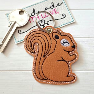 Könnte beinhalten: Ein brauner Leder-Schlüsselanhänger in Form eines Eichhörnchens mit schwarzen Stickdetails und einem Metallring. Ein silberner Schlüssel und ein Anhänger mit der Aufschrift "made with love" sind ebenfalls vorhanden.