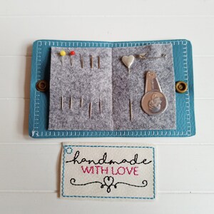 May include: A teal blue leather sewing kit with a gray felt interior. The kit contains needles, pins, a heart-shaped pincushion, a thimble, and a small sewing gauge. The kit has a white label with the text "handmade with love" in black and pink lettering.