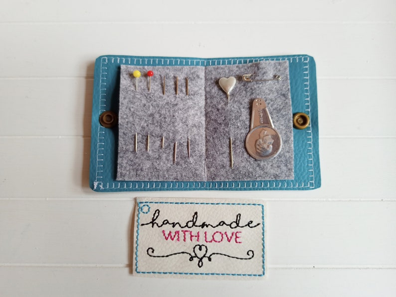 May include: A teal blue leather sewing kit with a gray felt interior. The kit contains needles, pins, a heart-shaped pincushion, a thimble, and a small sewing gauge. The kit has a white label with the text "handmade with love" in black and pink lettering.