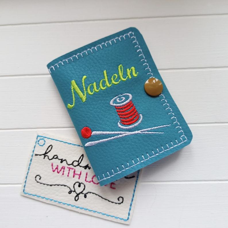 May include: A teal blue leather needle case with white stitching. The case has an embroidered design of a spool of red thread and a needle. The case has the word "Nadeln" embroidered on the front in green. A small brown button is on the front of the case. A white tag with black stitching says "handmade with love" in black and pink.