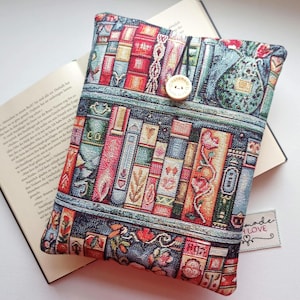 Könnte beinhalten: Ein farbenfrohes Buchcover mit einem Tapisserie-Design eines Bücherregals voller Bücher. Die Hülle, mit einem Knopf verschlossen, liegt über einem geöffneten Buch. Die Farbpalette umfasst Rot-, Blau- und Grüntöne.
