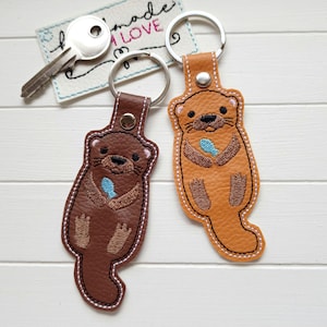 Könnte beinhalten: Zwei bestickte Otter-Schlüsselanhänger aus strukturiertem Kunstleder. Einer ist dunkelbraun und der andere hellbraun. Jeder Otter hält einen kleinen blauen Fisch und ist mit einer silbernen Niete an einem silbernen Metallring befestigt. Ein Schlüssel liegt bei.