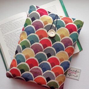 Könnte beinhalten: Ein farbenfroher Buchumschlag mit einem sich wiederholenden Muster aus sich überlappenden Halbkreisen in Rot-, Orange-, Gelb-, Grün-, Blau- und Lilatönen. Die Hülle hat einen Knopfverschluss und liegt neben einem offenen Buch.