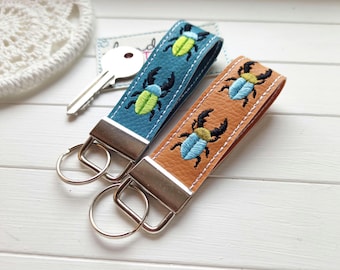 May include: Two leather keychains with silver metal hardware and split rings. One keychain is blue with embroidered lime green beetles, and the other is tan with embroidered light blue beetles. Both feature white stitching along the edges.
