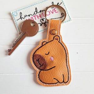 Könnte beinhalten: Ein brauner Leder-Schlüsselanhänger mit einem niedlichen gestickten Bären-Design. Der Bär hat geschlossene Augen und eine rosa Nase. Der Schlüsselanhänger ist an einem silbernen Schlüsselring befestigt.
