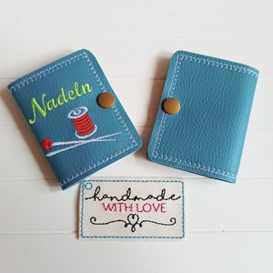 May include: Two blue leather needle cases with a gold button closure. One case has the word "Nadeln" embroidered on it with a red and white spool of thread and two needles. The other case is plain. A white tag with the text "handmade with love" is in the foreground.