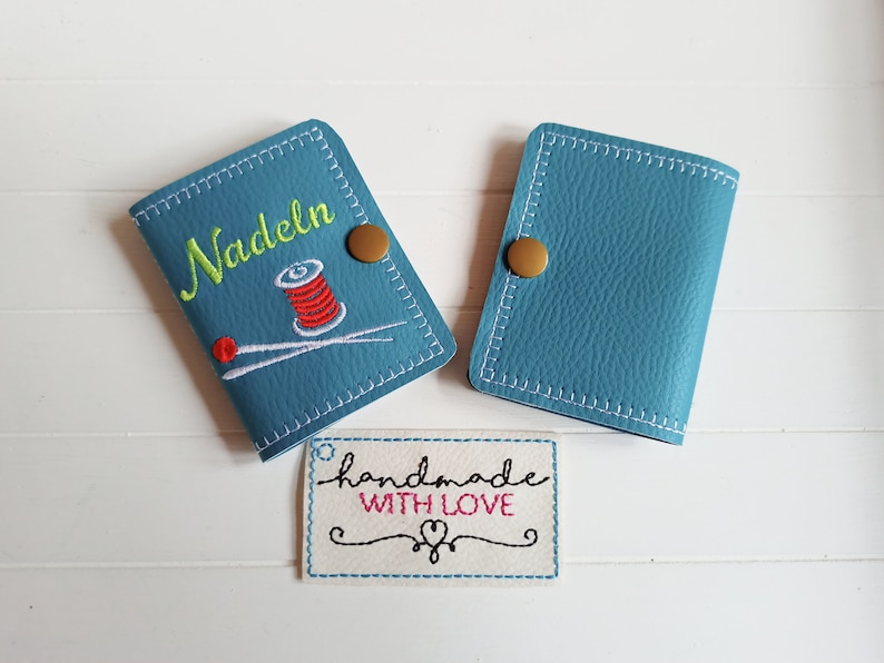 May include: Two blue leather needle cases with a gold button closure. One case has the word "Nadeln" embroidered on it with a red and white spool of thread and two needles. The other case is plain. A white tag with the text "handmade with love" is in the foreground.