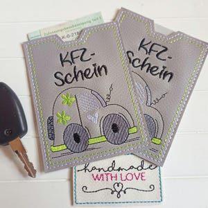 Könnte beinhalten: Zwei graue Stoff-Fahrzeugschein-Hüllen mit einem gestickten Design eines Autos und dem Text "KFZ-Schein" und "handgefertigt mit Liebe".