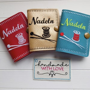 May include: Three small needle cases with embroidered designs. The cases are red, gold, and blue. Each case has the word "Nadeln" embroidered on the front. The gold case has a spool of thread and two needles embroidered on it. The red case has a spool of thread and two needles embroidered on it. The blue case has a spool of thread and two needles embroidered on it. A small white tag with the text "handmade with love" is in the foreground.