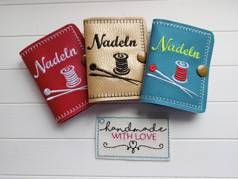May include: Three small needle cases with embroidered designs. The cases are red, gold, and blue. Each case has the word "Nadeln" embroidered on the front. The gold case has a spool of thread and two needles embroidered on it. The red case has a spool of thread and two needles embroidered on it. The blue case has a spool of thread and two needles embroidered on it. A small white tag with the text "handmade with love" is in the foreground.