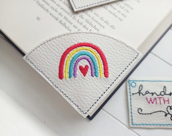 May include: White leather bookmark with an embroidered rainbow and a heart in the center. The bookmark is handmade and has a tag that says "handmade with love".