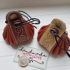 Könnte beinhalten: Zwei kleine, braune Lederbeutel mit dekorativen Nähten und Kordelzügen. Die Beutel haben einen hellbraunen Lederakzent mit gestickten Wirbeln. Eine kleine Karte mit dem Text "handmade WITH LOVE" und eine Münze sind ebenfalls im Bild.