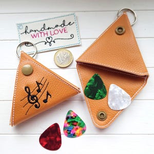 Könnte beinhalten: Zwei hellbraune, dreieckige Schlüsselanhänger aus Leder mit Metallringen. Einer hat einen schwarzen, gestickten Notenschlüssel und Noten. Der andere enthält drei Gitarrenplektren. Ein kleines gesticktes Etikett lautet "handmade WITH LOVE".