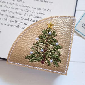 Könnte beinhalten: Goldfarbener Leder-Lesezeichen mit gesticktem Weihnachtsbaum-Design. Der Baum ist grün mit weißen und blauen Ornamenten und einem goldenen Stern. Das Lesezeichen liegt auf einem Buch.