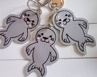 Puede incluir: Tres llaveros de fieltro gris con forma de focas con ojos, narices y bigotes bordados en negro. Cada foca tiene mejillas bordadas en rosa y está unida a un llavero plateado.