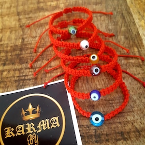 Könnte beinhalten: Fünf rote Armbänder mit blauen, grünen, orangen, weißen und violetten bösen Augen-Anhängern. Die Armbänder sind übereinander gestapelt. Eine weiße Karte mit dem Text "KARMA BY Natalia" befindet sich im Hintergrund.