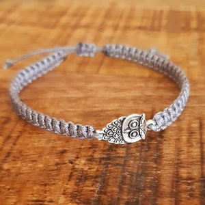 Puede incluir: Un brazalete trenzado gris con un dije de búho plateado.