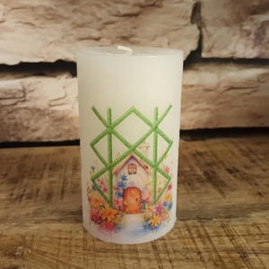 Könnte beinhalten: Weiße Kerze in Säulenform mit einem grün-weißen Design eines Hauses mit einer Tür und Blumen. Das Design ist von einem grünen geometrischen Muster umgeben.