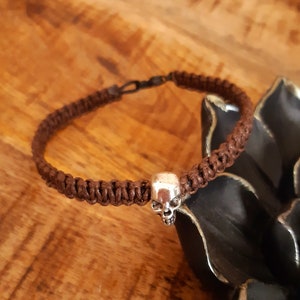 Könnte beinhalten: Ein braunes geflochtenes Armband mit einem silbernen Totenkopf-Anhänger.