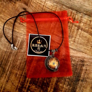 May include: A brown leather cord necklace with a gold-toned pendant featuring an Om symbol inside a circular design. The necklace is packaged in a red organza bag with a white tag that reads "Karma by Natalie".